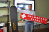 a man holding a metal snowboard patrol sign with a red background and white text that reads 'Snowboard Patrol' in one hand and a snowboard in the other hand