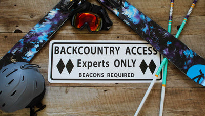 metal sign with black text saying 'backcountry access experts only beacon required' with a white background and various ski gear around the sign