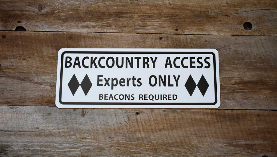 metal sign with black text saying 'backcountry access experts only beacon required' with a white background and various ski gear around the sign