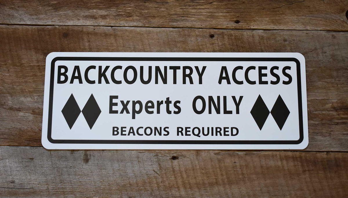 metal sign with black text saying 'backcountry access experts only beacon required' with a white background on a wood background