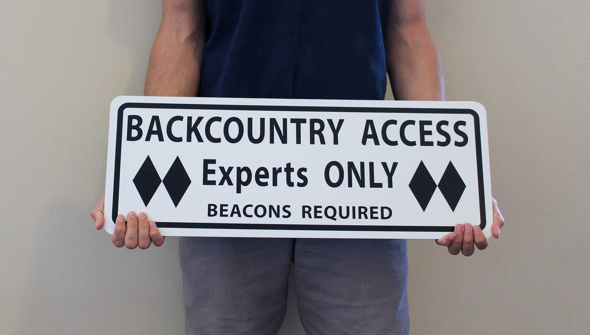man holding a metal sign with black text saying 'backcountry access experts only beacon required' with a white background