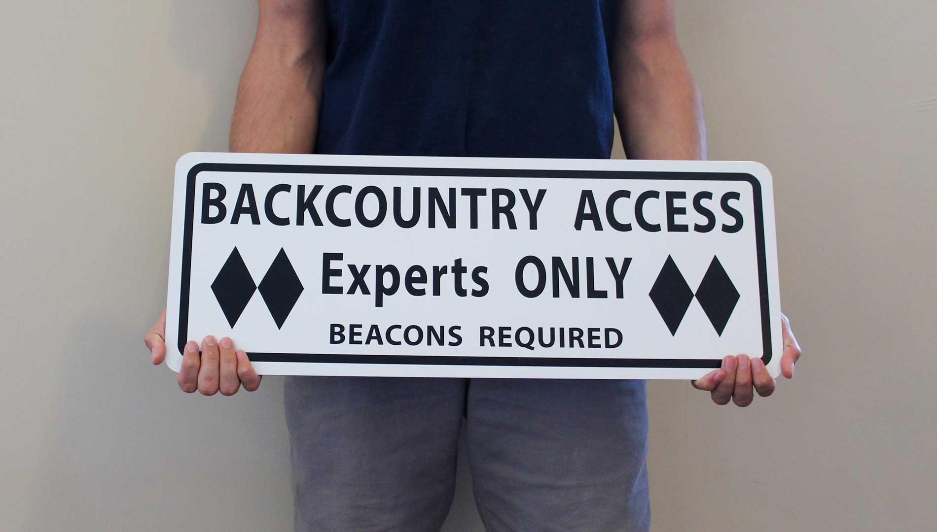 man holding a metal sign with black text saying 'backcountry access experts only beacon required' with a white background