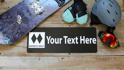 metal ski run sign with a black background and white text that says 'Your Text Here' with two black diamonds surrounded by ski gear