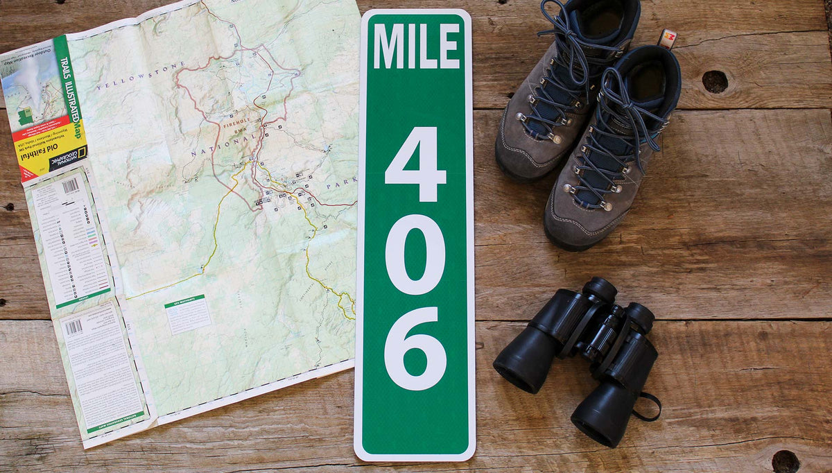 custom metal mile marker sign with a green background and white lettering with travel gear around the sign
