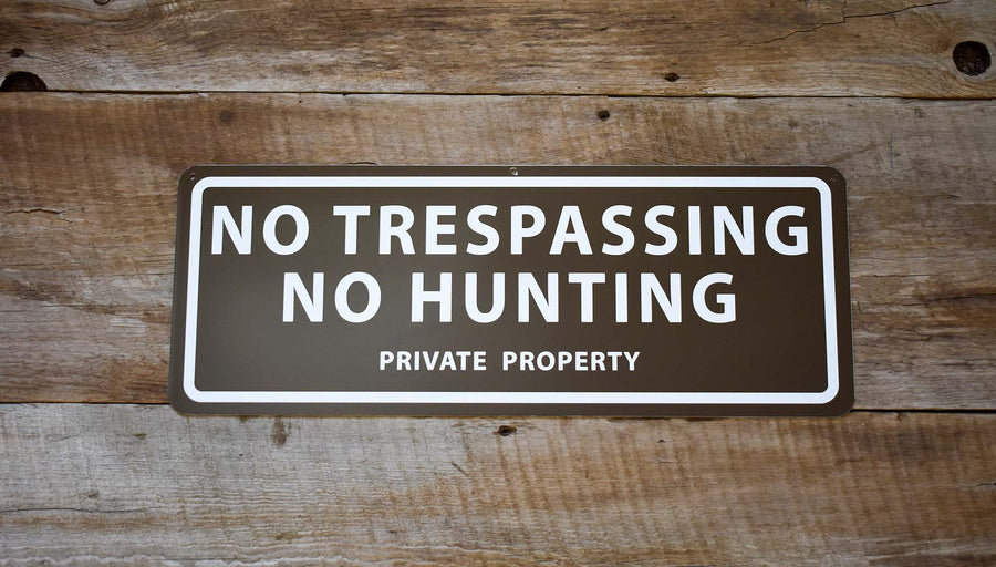 metal sign with a brown background and white text saying 'no trespassing no hunting private property' with antlers and pine needles around the sign