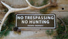 metal sign with a brown background and white text saying 'no trespassing no hunting private property' with antlers and pine needles around the sign
