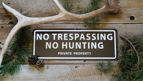 metal sign with a brown background and white text saying 'no trespassing no hunting private property' with antlers and pine needles around the sign
