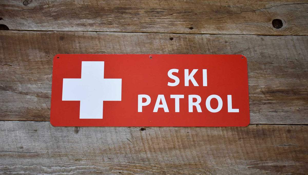 a metal ski patrol sign with a red background and white text that reads 'Ski Patrol' on a wooden background
