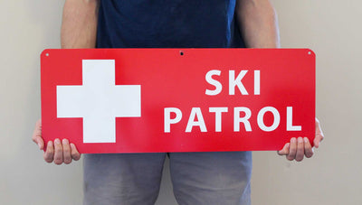 a man holding a metal ski patrol sign with a red background and white text that reads 'Ski Patrol'