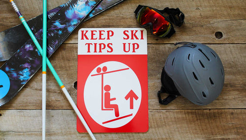 a metal sign with a white background and red text that says 'Keep Ski Tips Up' with an image of a skier on a chairlift surrounded by ski gear