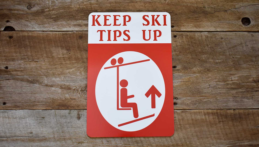 a metal sign with a white background and red text that says 'Keep Ski Tips Up' with an image of a skier on a chairlift surrounded by ski gear