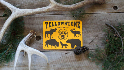 metal sign with a yellow background and black text that says 'Yellowstone Big 5' with black outlines of a moose, elk, bison, wolf, and grizzly bear with antlers and pine needles and cones around it