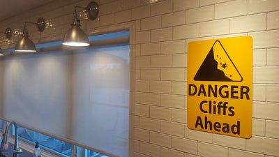 metal sign with black text saying 'danger cliffs ahead' with a yellow background and image of falling rock hung up on a wall