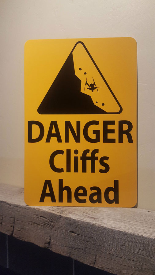 close up picture of metal sign with black text saying 'danger cliffs ahead' with a yellow background and image of falling rock