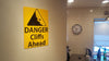angled photo of a metal sign with black text saying 'danger cliffs ahead' with a yellow background and image of falling rock