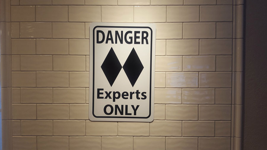 metal sign with black text saying 'danger experts only' with a two black diamonds on a white background hung up on a wall