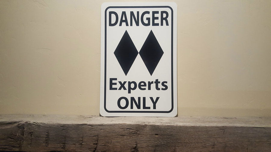 metal sign with black text saying 'danger experts only' with a two black diamonds on a white background on a mantle