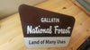 brown and white metal sign with text saying 'gallatin national forest land of many uses' on a wooden background