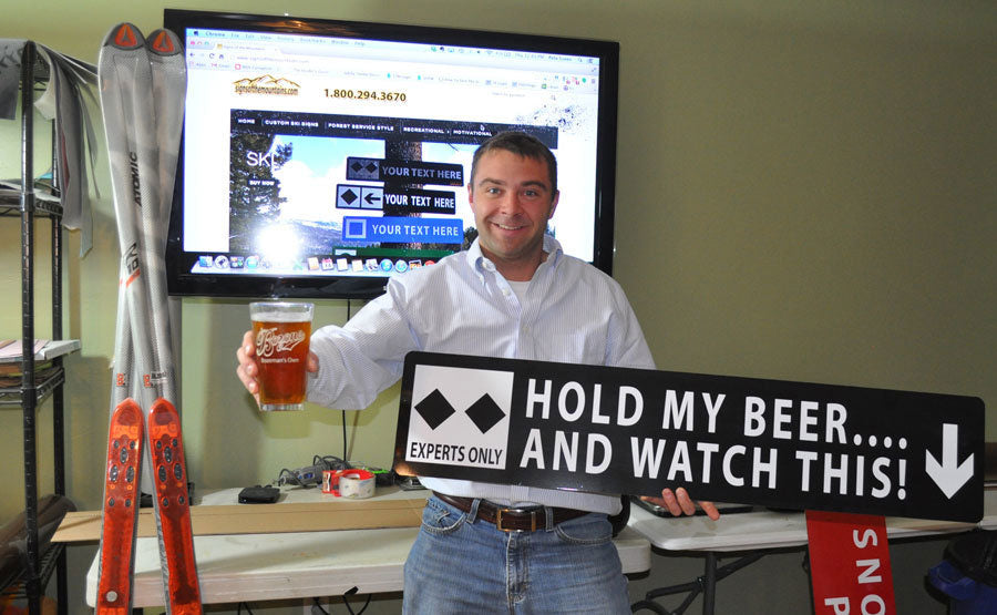 man holding up a beer and a metal sign with white text saying 'hold my beer...and watch this!' with a black background and two black diamonds