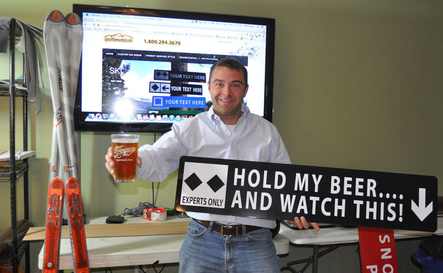 metal sign with white text saying 'hold my beer...and watch this!' with a black background and two black diamonds
