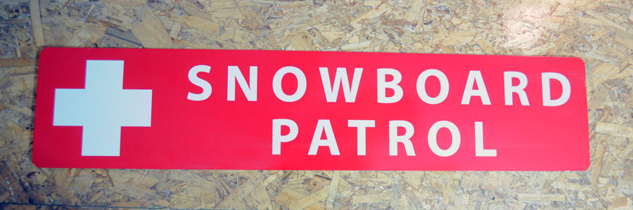 a metal snowboard patrol sign with a red background and white text that reads 'Snowboard Patrol' hung up on a wall