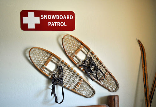 a metal snowboard patrol sign with a red background and white text that reads 'Snowboard Patrol' hung up on a wall