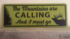 metal sign with a yellow background and black text saying 'the mountains are calling and i must go' with a black elk and tree outline