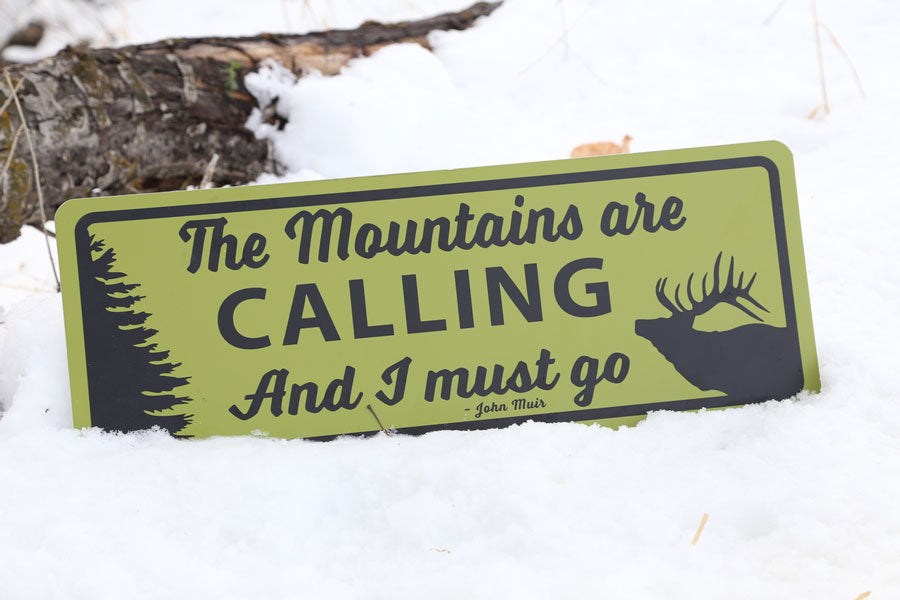 metal sign with a yellow background and black text saying 'the mountains are calling and i must go' in the snow